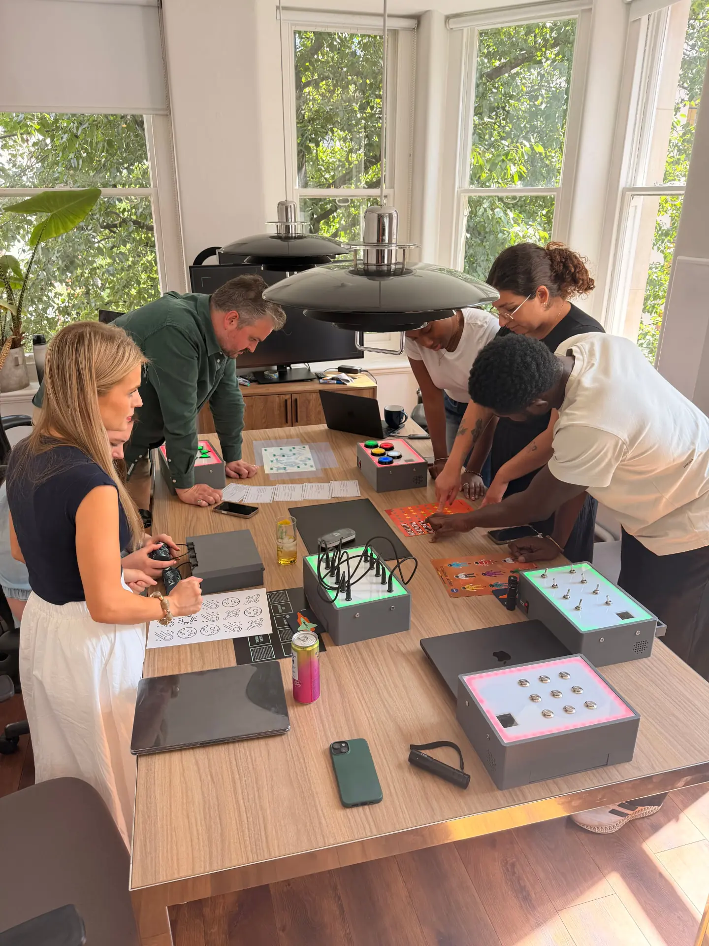 A team leaning over a meeting room table working through an UNPACK session, with lit puzzle modules in red and green, mission documents and physical puzzles in play.